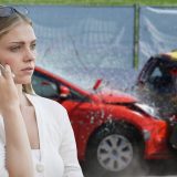 Wystraszona kobieta rozmawia przez telefon, z tyłu widać wypadek samochodowy, dwa samochody zderzone ze sobą czołowo